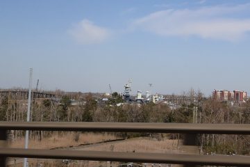 Distant view of Battleship North Carolina beyond trees and a riverbank in Wilmington, seen past a highway guardrail