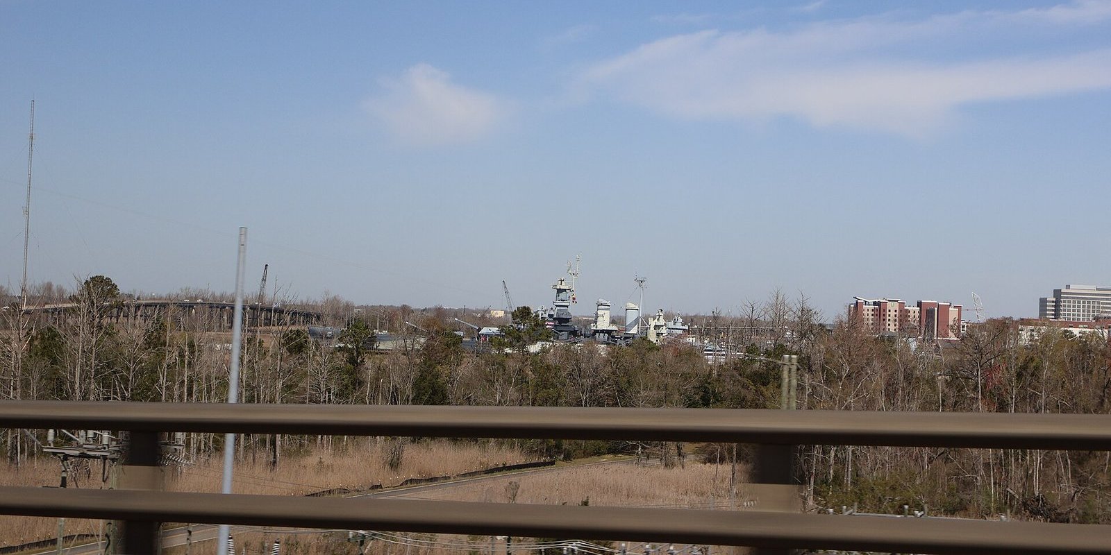 Distant view of Battleship North Carolina beyond trees and a riverbank in Wilmington, seen past a highway guardrail