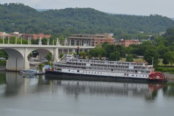 White Delta Queen sternwheel riverboat with a red paddlewheel docked on the Tennessee River in Chattanooga near an arched bridge