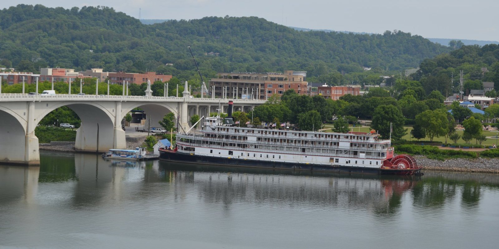 White Delta Queen sternwheel riverboat with a red paddlewheel docked on the Tennessee River in Chattanooga near an arched bridge