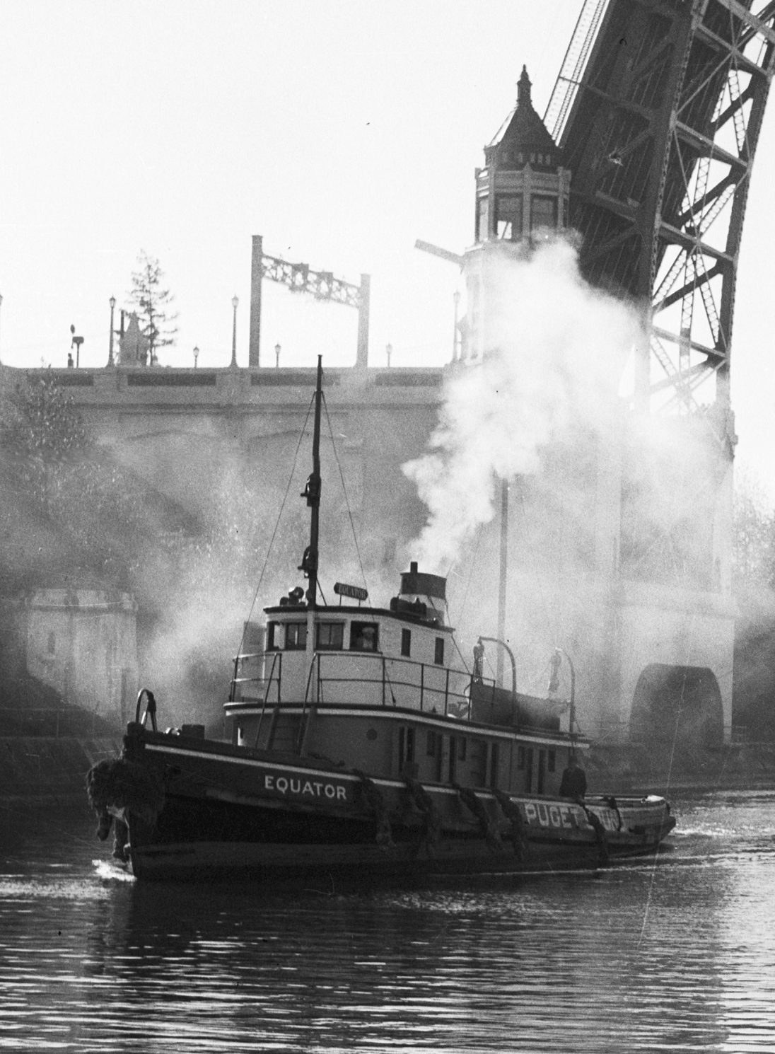 Black-and-white photograph of the tugboat Equator passing through the Montlake Cut in Seattle with the Montlake Bridge behind