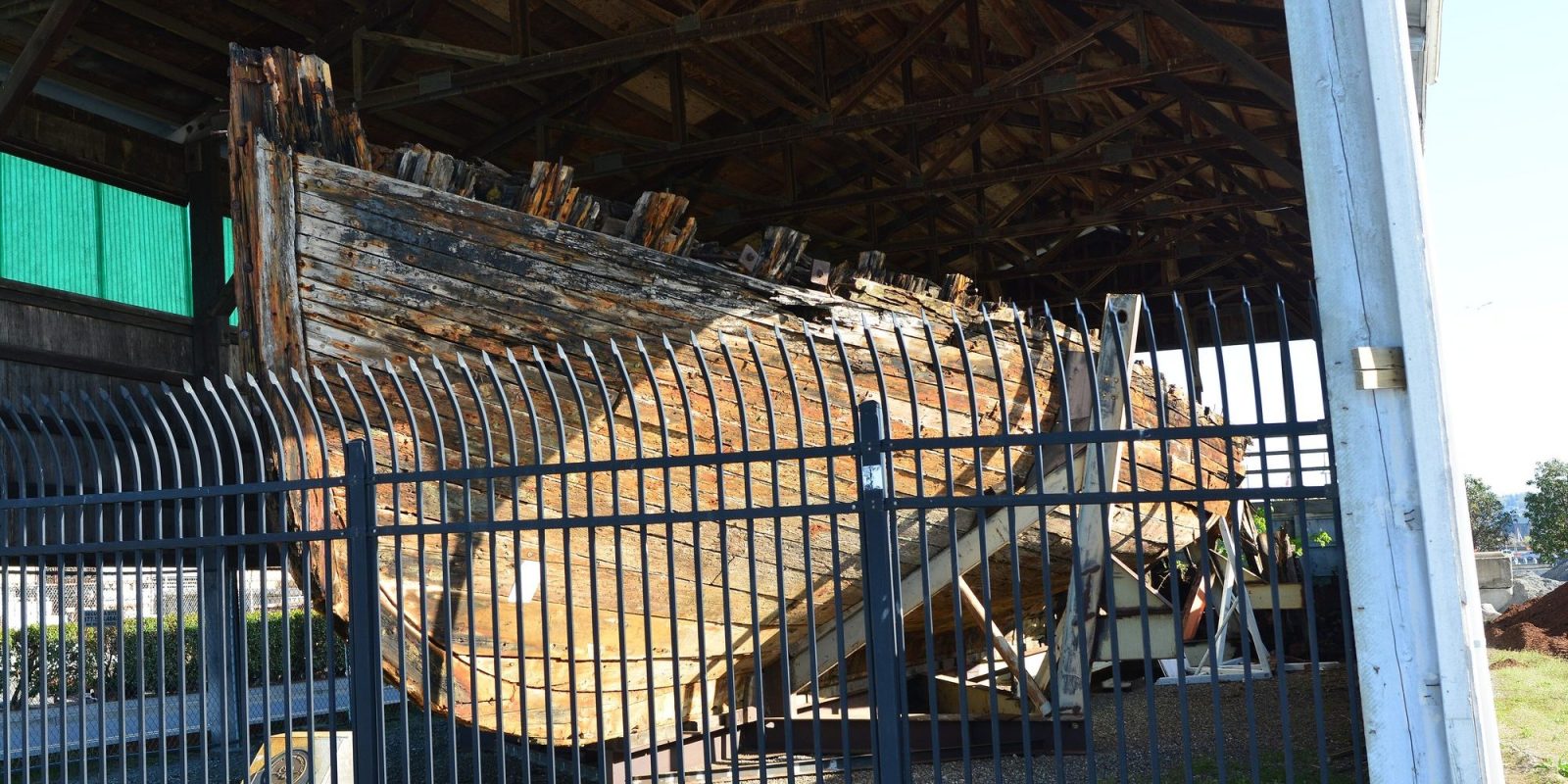 The ruined wooden hull of the schooner Equator on blocks inside an open shed behind a metal fence in Everett, Washington