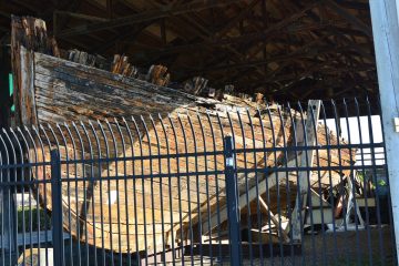 The ruined wooden hull of the schooner Equator on blocks inside an open shed behind a metal fence in Everett, Washington