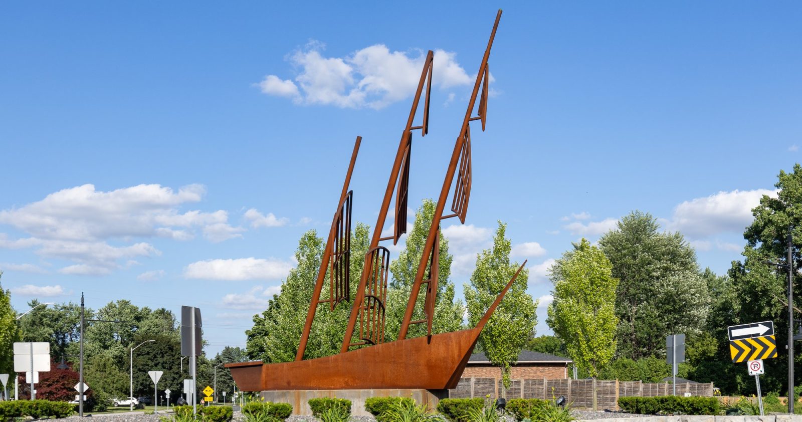 A rust-colored steel sculpture of a three-masted sailing ship standing on a stone base in a landscaped roundabout under a blue sky