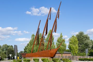 A rust-colored steel sculpture of a three-masted sailing ship standing on a stone base in a landscaped roundabout under a blue sky