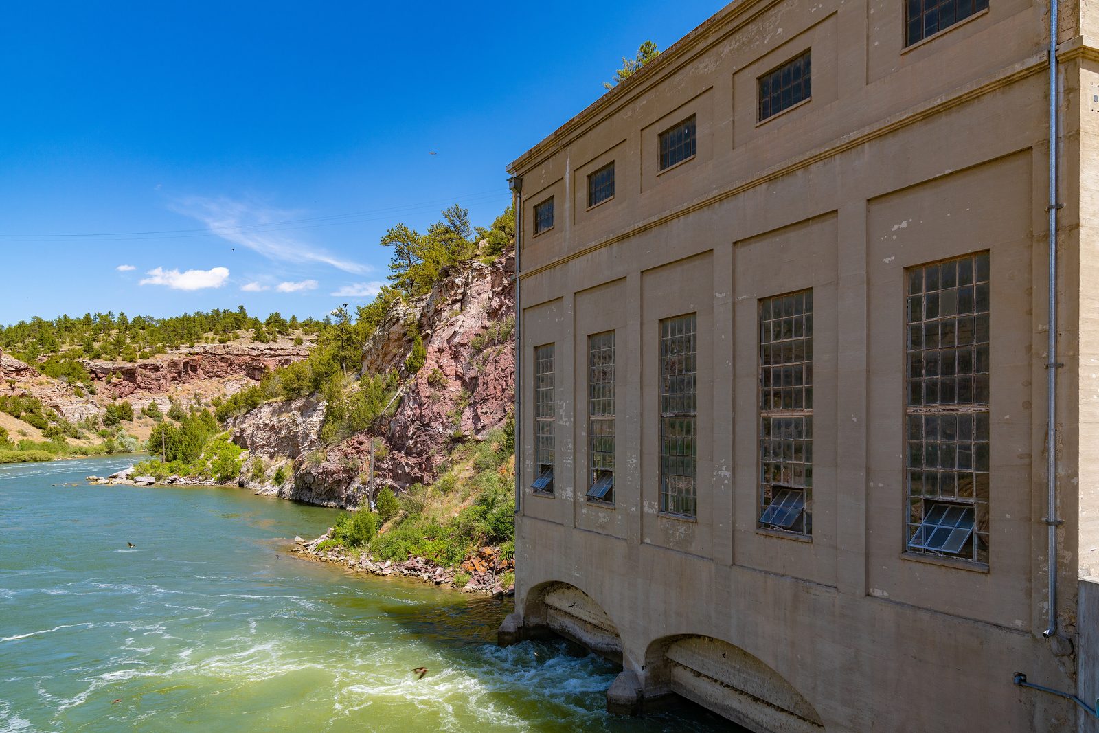 The North Platte River flowing past the concrete power plant at Guernsey Dam, between pink rock cliffs under a blue sky