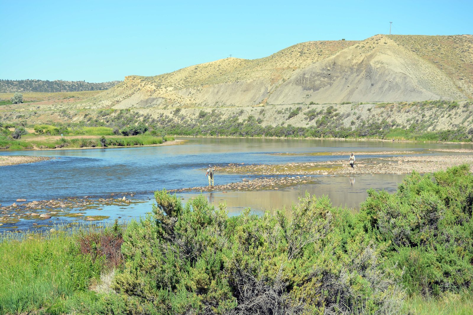 Two anglers wading across gravel bars in the wide, shallow North Platte River below sagebrush hills