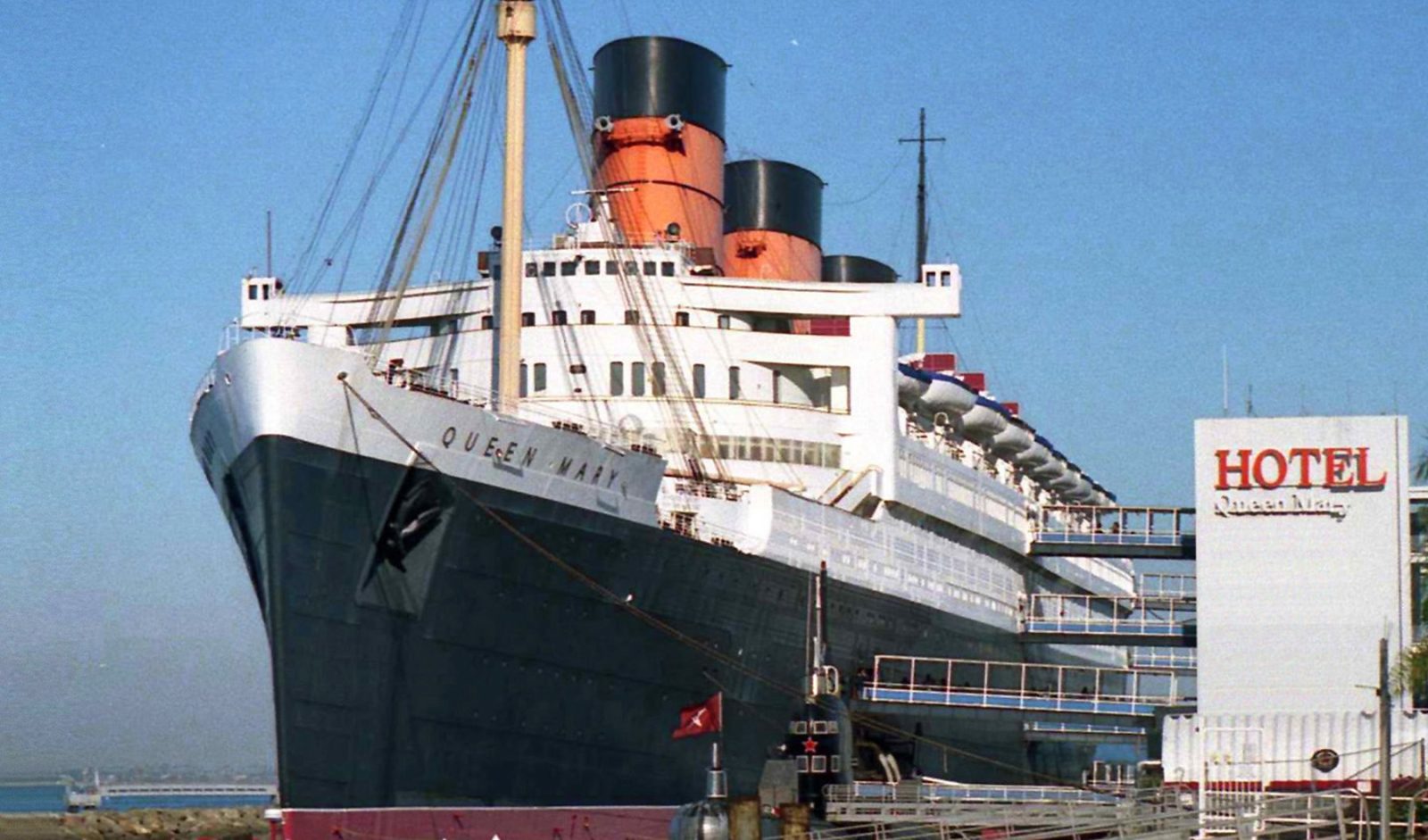 Ocean liner Queen Mary with black hull and red-and-black funnels docked in Long Beach beside a Hotel sign