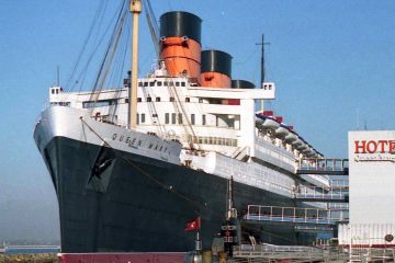 Ocean liner Queen Mary with black hull and red-and-black funnels docked in Long Beach beside a Hotel sign