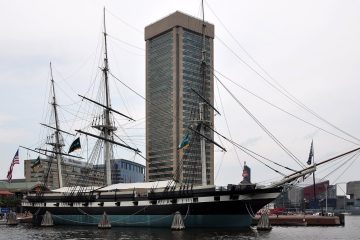 Sloop-of-war USS Constellation with tall masts and rigging moored in Baltimore Inner Harbor beside a high-rise tower