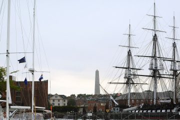 Three-masted USS Constitution docked in Boston with the Bunker Hill Monument behind and sailboat masts in the foreground