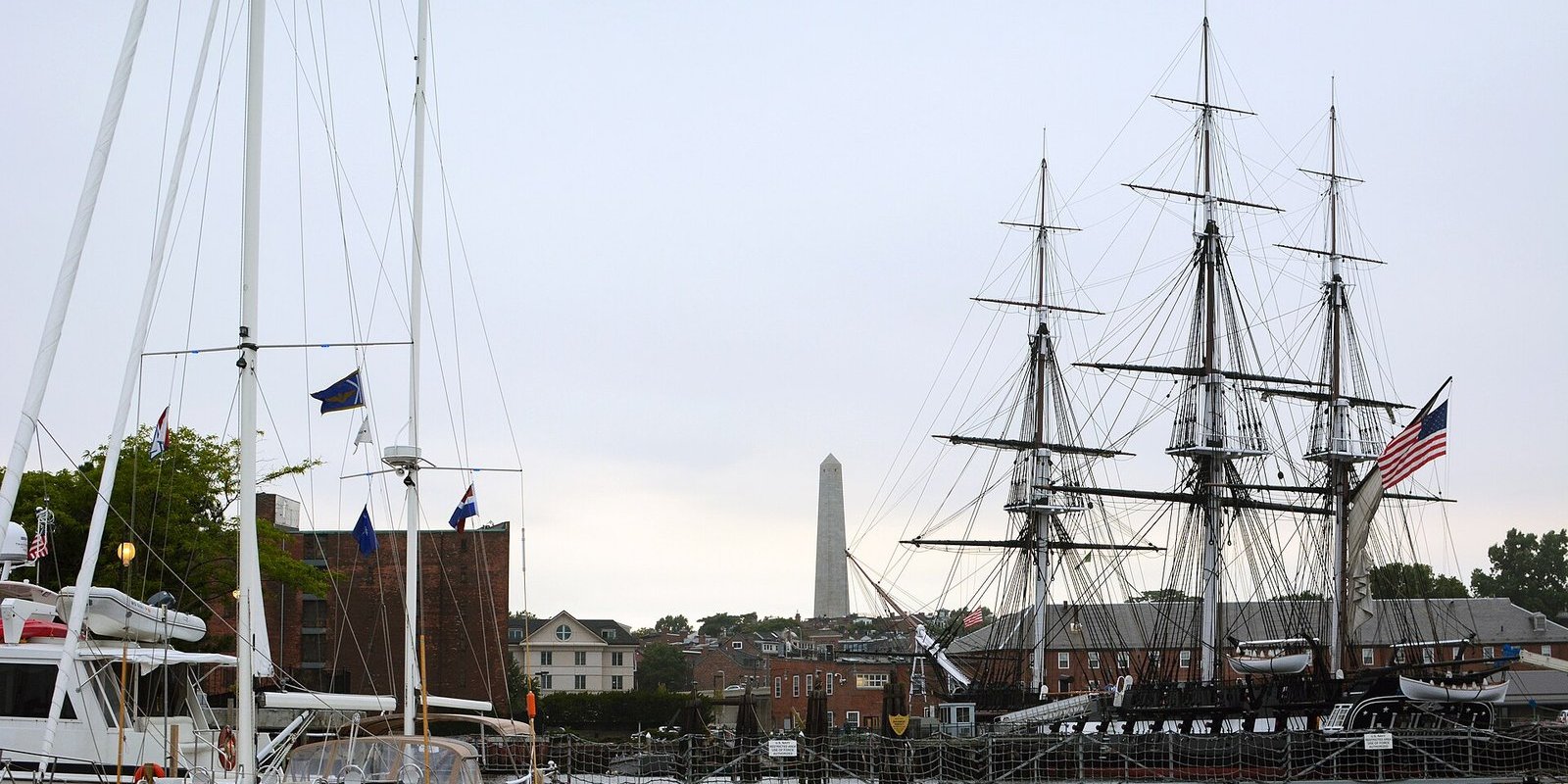 Three-masted USS Constitution docked in Boston with the Bunker Hill Monument behind and sailboat masts in the foreground