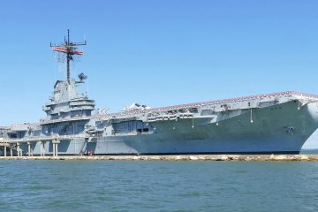 Gray aircraft carrier USS Lexington moored along a seawall in Corpus Christi Bay under a blue sky