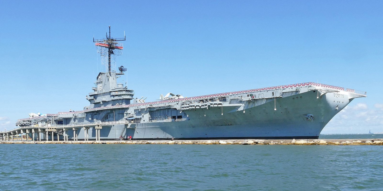 Gray aircraft carrier USS Lexington moored along a seawall in Corpus Christi Bay under a blue sky